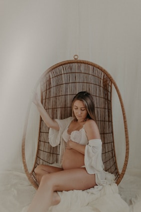 A pregnant woman sits comfortably in a woven hanging chair with a soft, serene expression. She is dressed in a white lace bra and an open robe, gently cradling her belly. The setting exudes a calm, intimate atmosphere with neutral and warm tones.