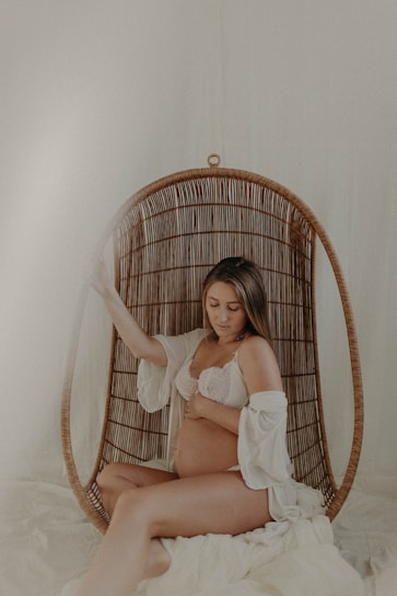 A pregnant woman sits comfortably in a woven hanging chair with a soft, serene expression. She is dressed in a white lace bra and an open robe, gently cradling her belly. The setting exudes a calm, intimate atmosphere with neutral and warm tones.