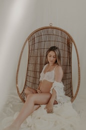 A woman is seated in a wicker hanging chair, gently cradling her pregnant belly. She wears a white lingerie set and a lightly draped white fabric, with a serene, introspective expression on her face. The setting is softly lit, creating a calm and intimate atmosphere.