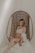 A woman is seated in a wicker hanging chair, gently cradling her pregnant belly. She wears a white lingerie set and a lightly draped white fabric, with a serene, introspective expression on her face. The setting is softly lit, creating a calm and intimate atmosphere.