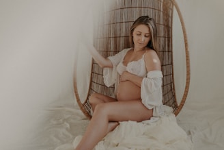 A pregnant woman is seated on a wicker chair with a serene expression. She wears a white lace bralette and a matching sheer robe, gently holding her stomach. The setting has a soft, muted ambiance with cream and beige tones, adding to the overall tranquility.
