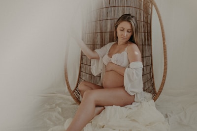 A pregnant woman is seated on a wicker chair with a serene expression. She wears a white lace bralette and a matching sheer robe, gently holding her stomach. The setting has a soft, muted ambiance with cream and beige tones, adding to the overall tranquility.