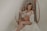 A pregnant woman is seated on a wicker chair with a serene expression. She wears a white lace bralette and a matching sheer robe, gently holding her stomach. The setting has a soft, muted ambiance with cream and beige tones, adding to the overall tranquility.