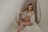 A pregnant woman is seated on a wicker chair with a serene expression. She wears a white lace bralette and a matching sheer robe, gently holding her stomach. The setting has a soft, muted ambiance with cream and beige tones, adding to the overall tranquility.