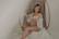 A pregnant woman is seated on a wicker chair with a serene expression. She wears a white lace bralette and a matching sheer robe, gently holding her stomach. The setting has a soft, muted ambiance with cream and beige tones, adding to the overall tranquility.