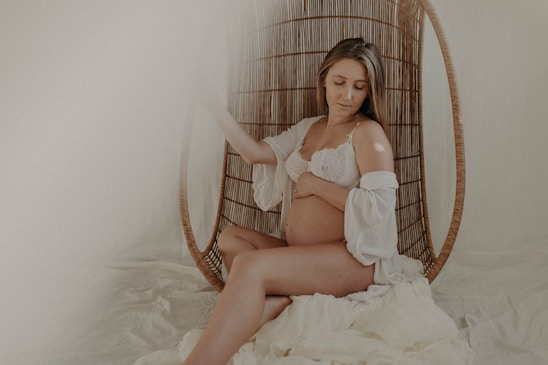 A pregnant woman is seated on a wicker chair with a serene expression. She wears a white lace bralette and a matching sheer robe, gently holding her stomach. The setting has a soft, muted ambiance with cream and beige tones, adding to the overall tranquility.