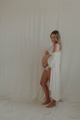 A pregnant woman stands barefoot on a soft, light-colored surface, gently cradling her belly. She wears a flowing white garment that drapes elegantly over her body. Her expression is serene, and she looks downwards, lost in thought. The background is a plain, soft white, enhancing the peaceful and intimate atmosphere of the scene.