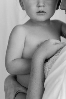 A black and white photograph featuring a close-up of a young child being held gently. The child is unclothed, and an adult's arm, covered by lace fabric, supports the child. The image focuses on the bond and intimacy between the two.