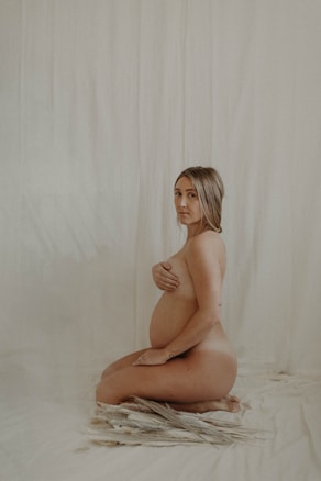 A nude pregnant woman kneels on a soft fabric backdrop, with one hand gently holding her belly and the other covering her chest. The setting is minimalistic, with a neutral color palette.