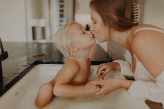 A mother and child share a tender moment in a kitchen setting. The child is sitting in a sink filled with water, while the mother leans over to give the child a gentle kiss on the lips. The soft lighting and serene expressions highlight the intimacy and warmth of the scene.