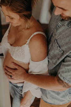 A couple is standing closely together, with the man's hand gently placed on the woman's bare abdomen. The woman is wearing a white lace top, and the man has a tattoo on his arm. They both appear to be smiling or looking at the woman's belly, suggesting a moment of connection or tenderness.