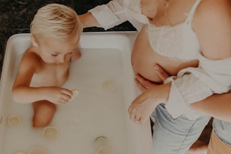 A young child is bathing in a white tub filled with milky water containing slices of what appears to be lemon or a similar fruit. An adult, possibly a pregnant woman, stands nearby, with one hand resting on her baby bump and the other gently touching the child's arm.