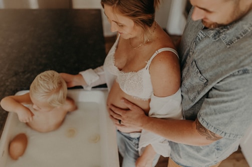 A family moment with a child taking a bath in a small tub filled with water, as a man and a pregnant woman gently touch and watch over him. The woman wears a white lace top and the man has a denim shirt, showcasing an intimate and caring atmosphere.