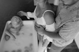 A child with light hair is sitting in a bathtub filled with milk or water, accompanied by slices of citrus floating around. Next to the tub, two adults, one of whom appears pregnant, are standing with their hands on the woman's belly. The scene is captured in black and white, giving it a timeless and intimate feel.