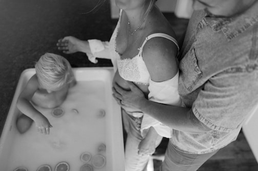 A child with light hair is sitting in a bathtub filled with milk or water, accompanied by slices of citrus floating around. Next to the tub, two adults, one of whom appears pregnant, are standing with their hands on the woman's belly. The scene is captured in black and white, giving it a timeless and intimate feel.