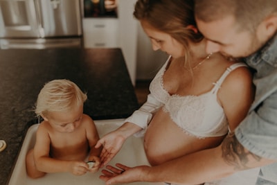 A pregnant woman stands with a man and a small child who is playing with a toy egg or shell. The scene takes place in a kitchen setting with a counter and appliances visible. The woman wears a white bra, and the man is leaning in closely, appearing supportive.