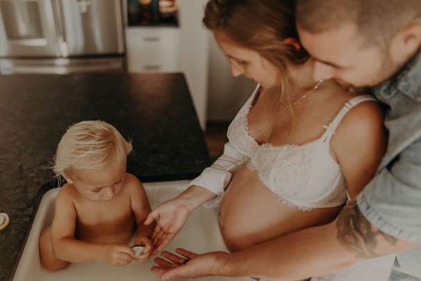 A pregnant woman stands with a man and a small child who is playing with a toy egg or shell. The scene takes place in a kitchen setting with a counter and appliances visible. The woman wears a white bra, and the man is leaning in closely, appearing supportive.