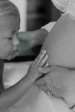 A tender moment featuring a young child gently touching a woman's pregnant belly. The child's eyes are closed, and their hands rest softly upon the belly, which is partially covered with lace fabric.