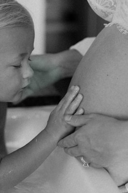 A tender moment featuring a young child gently touching a woman's pregnant belly. The child's eyes are closed, and their hands rest softly upon the belly, which is partially covered with lace fabric.