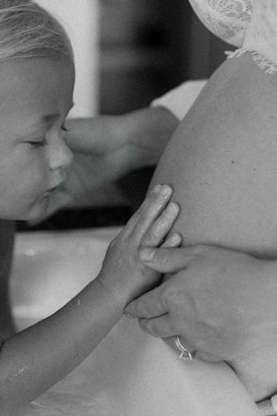 A tender moment featuring a young child gently touching a woman's pregnant belly. The child's eyes are closed, and their hands rest softly upon the belly, which is partially covered with lace fabric.
