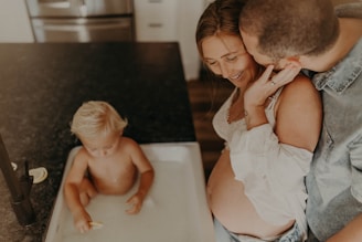 A pregnant woman is affectionately embraced by a man while smiling at a young child playing in water inside a sink. The setting appears to be a home kitchen with a countertop and faucet visible.