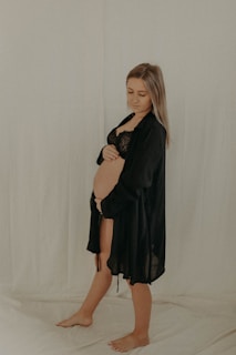 A woman stands barefoot on a draped surface. She is wearing a black lace bra and a black robe that is slightly open, revealing her pregnant belly. Her right hand rests gently on her stomach while she looks down with a calm expression. The background is a soft, light color, creating a serene atmosphere.