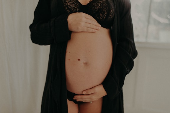 A pregnant woman is gently holding her bare belly with both hands. She is wearing a black lacy bra and an open black robe. The background is softly blurred, with a light, neutral tone.