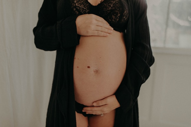 A pregnant woman is gently holding her bare belly with both hands. She is wearing a black lacy bra and an open black robe. The background is softly blurred, with a light, neutral tone.