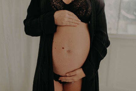A pregnant woman is gently holding her bare belly with both hands. She is wearing a black lacy bra and an open black robe. The background is softly blurred, with a light, neutral tone.