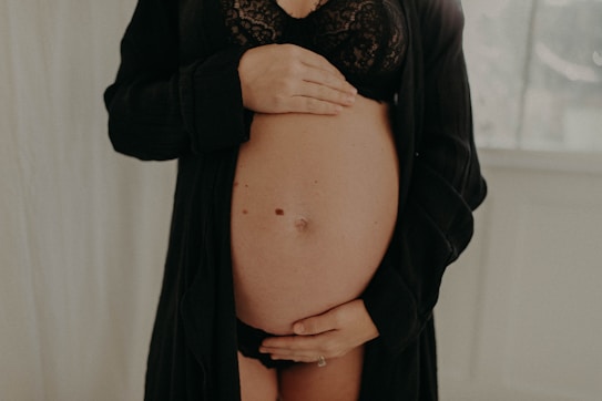 A pregnant woman is gently holding her bare belly with both hands. She is wearing a black lacy bra and an open black robe. The background is softly blurred, with a light, neutral tone.