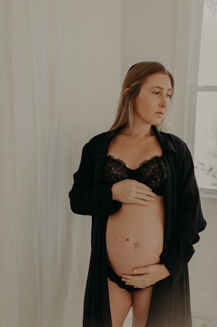 A pregnant woman wearing black lingerie and a loose robe is standing in a softly lit room, gently resting her hands on her belly. She appears contemplative and serene, with natural light coming through a window on one side.