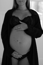 A black and white photograph of a pregnant woman. She is dressed in lingerie and a robe, with one hand resting on her upper belly and the other hand positioned below her belly. A ring is visible on one of her fingers. The background is softly blurred, emphasizing her figure.
