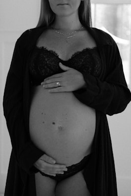 A black and white photograph of a pregnant woman. She is dressed in lingerie and a robe, with one hand resting on her upper belly and the other hand positioned below her belly. A ring is visible on one of her fingers. The background is softly blurred, emphasizing her figure.