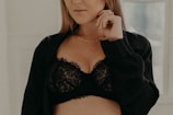 A person is wearing a black lace bra with a black shirt loosely draped over. They are holding their hand near their face, adorned with rings and a delicate necklace.