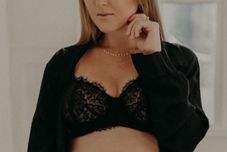 A person is wearing a black lace bra with a black shirt loosely draped over. They are holding their hand near their face, adorned with rings and a delicate necklace.