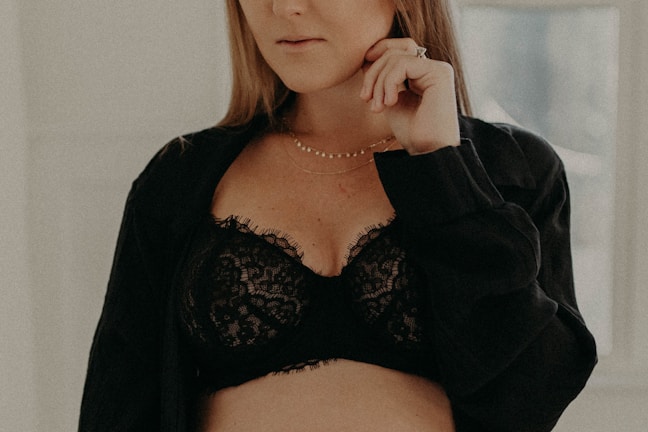 A person is wearing a black lace bra with a black shirt loosely draped over. They are holding their hand near their face, adorned with rings and a delicate necklace.