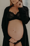 A pregnant woman gently cradles her belly with one hand while wearing black lace lingerie and a matching robe. The focus is on her midsection, and her face is partially visible, conveying a serene and introspective mood.
