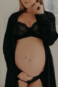 A pregnant woman gently cradles her belly with one hand while wearing black lace lingerie and a matching robe. The focus is on her midsection, and her face is partially visible, conveying a serene and introspective mood.