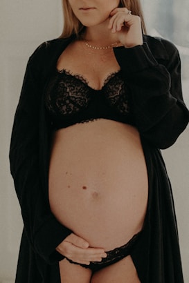 A pregnant woman gently cradles her belly with one hand while wearing black lace lingerie and a matching robe. The focus is on her midsection, and her face is partially visible, conveying a serene and introspective mood.