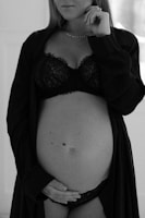 A black and white photo of a pregnant woman standing, wearing a lace bra and partially open robe. Her hand rests gently on her stomach, conveying a sense of calm and introspection.