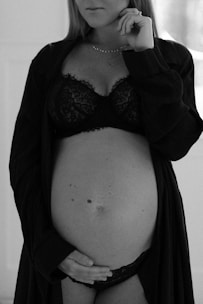 A black and white photo of a pregnant woman standing, wearing a lace bra and partially open robe. Her hand rests gently on her stomach, conveying a sense of calm and introspection.
