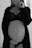 A black and white photo of a pregnant woman standing, wearing a lace bra and partially open robe. Her hand rests gently on her stomach, conveying a sense of calm and introspection.