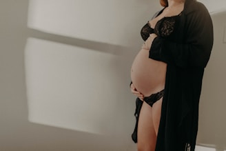A pregnant woman is dressed in black lingerie, cradling her belly with one hand. The soft lighting creates gentle shadows on the white wall behind her, adding a serene and intimate atmosphere.