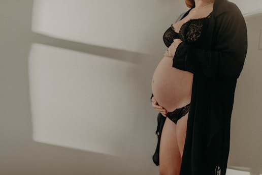 A pregnant woman is dressed in black lingerie, cradling her belly with one hand. The soft lighting creates gentle shadows on the white wall behind her, adding a serene and intimate atmosphere.
