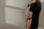 A pregnant woman is dressed in black lingerie, cradling her belly with one hand. The soft lighting creates gentle shadows on the white wall behind her, adding a serene and intimate atmosphere.