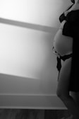 A monochrome photo of a pregnant person standing sideways, gently holding their bare belly. The person is wearing a dark bra and a draped garment. The light creates soft shadows on the wall and floor, adding depth and contrast.