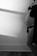 A monochrome photo of a pregnant person standing sideways, gently holding their bare belly. The person is wearing a dark bra and a draped garment. The light creates soft shadows on the wall and floor, adding depth and contrast.