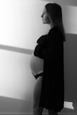 A black and white photo of a pregnant woman standing in profile with a soft light casting shadows on the wall. She is wearing a black lace bralette and an open shirt, gazing into the distance with a serene expression.
