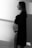 A black and white photo of a pregnant woman standing in profile with a soft light casting shadows on the wall. She is wearing a black lace bralette and an open shirt, gazing into the distance with a serene expression.