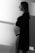 A black and white photo of a pregnant woman standing in profile with a soft light casting shadows on the wall. She is wearing a black lace bralette and an open shirt, gazing into the distance with a serene expression.